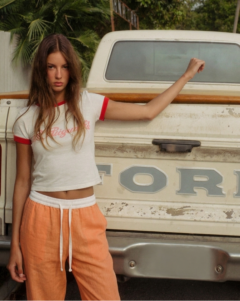 A woman stands in front of a vintage Ford pickup truck wearing a white ringer T-shirt with red trim and pink script lettering, paired with peach-orange drawstring sweatpants.